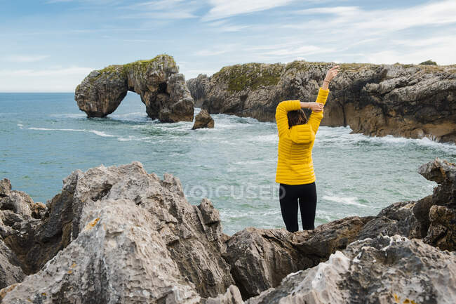 Back view unrecognizable female tourist in warm yellow jacket standing on rocky seashore and raising arms while admiring seascape views — Stock Photo