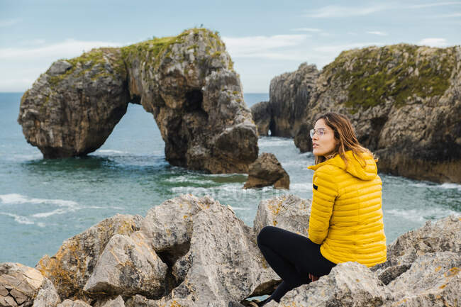 Visão traseira corpo inteiro feminino sereno em jaqueta quente sentado em pedras e admirando o mar azul acenando no dia de outono claro — Fotografia de Stock