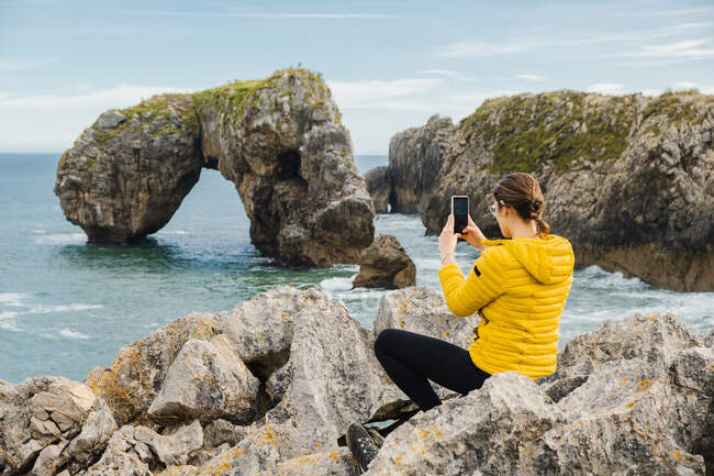 Vista posterior irreconocible viajero femenino en chaqueta de color amarillo cálido sentado en piedras afiladas y tomar fotos en el teléfono móvil de pintoresco paisaje marino - foto de stock