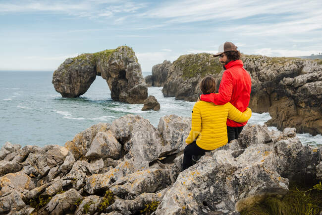 Back view young couple in warm jackets embracing and sitting on sharp stones on vast seacoast while admiring picturesque sea views — Stock Photo