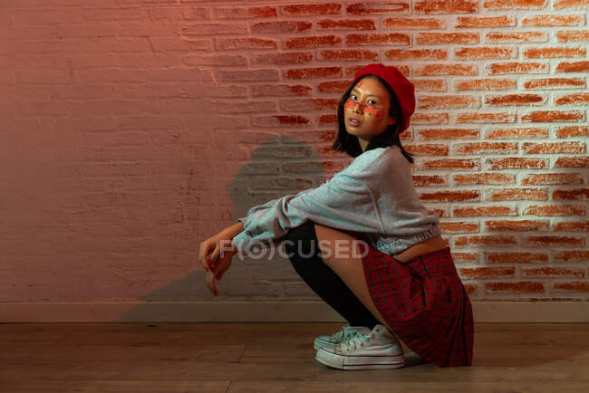 Pleine longueur confiant jeune asiatique femelle dans élégant tenue et chapeau assis sur parquet et regardant caméra dans chambre sombre contre mur de briques — Photo de stock