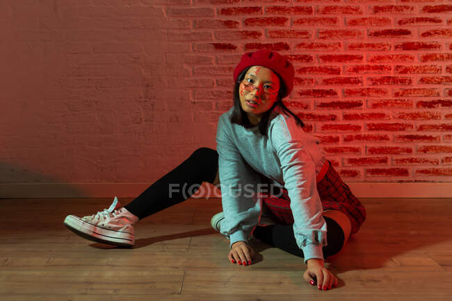 Piena lunghezza fiducioso giovane donna asiatica in abito elegante e cappello seduto sul parquet e guardando la fotocamera in camera oscura contro muro di mattoni — Foto stock