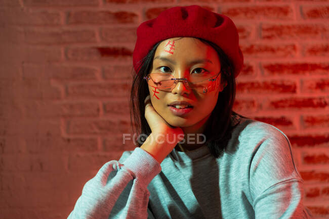 Уверенная молодая азиатка в стильной одежде и шляпе сидит на паркете и смотрит на камеру в темной комнате напротив кирпичной стены — стоковое фото