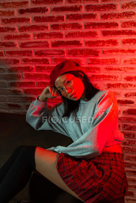Fiducioso giovane donna asiatica in abito elegante e cappello seduto sul parquet e guardando la fotocamera in camera oscura contro muro di mattoni — Foto stock