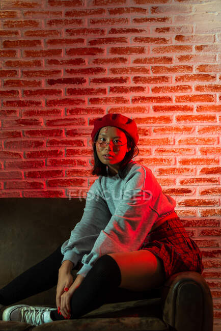 Цілковита молода азіатка в стильному одязі і капелюсі сидить на дивані і дивиться на камеру в темній кімнаті проти цегляної стіни. — стокове фото