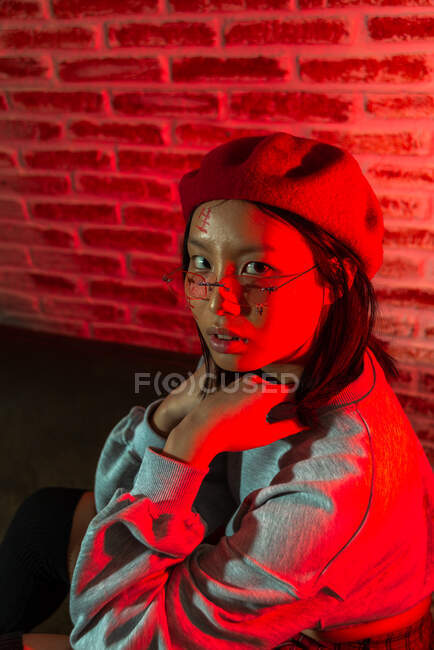 Selbstbewusste junge Asiatin in stylischem Outfit und Hut sitzt auf Parkett und blickt in dunklen Raum gegen Ziegelwand in die Kamera — Stockfoto