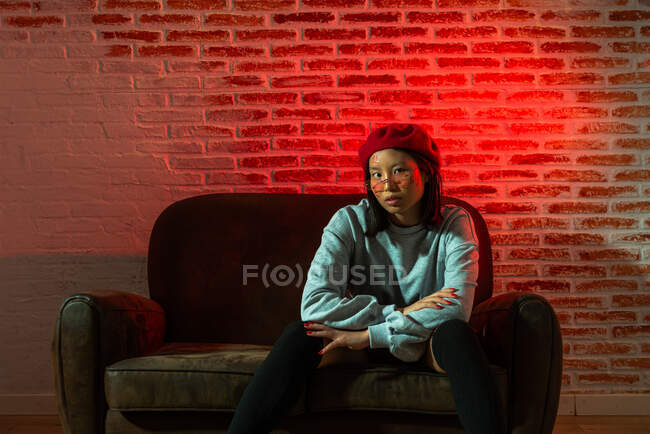 Comprimento total confiante jovem Asiática fêmea em roupa elegante e chapéu sentado no sofá e olhando para a câmera no quarto escuro contra a parede de tijolo — Fotografia de Stock