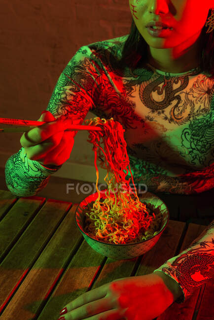 Jeune femme asiatique sans émotion avec des hiéroglyphes peints sur le visage portant un chapeau élégant mangeant de délicieuses nouilles tout en étant assis dans une pièce sombre contre un mur de briques — Photo de stock