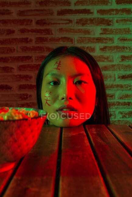 Unbewegte junge Asiatinnen mit Hieroglyphen auf hübschem Gesicht, angelehnt an Holztisch mit Nudel in Schale und Blick in die Kamera — Stockfoto