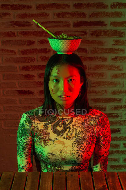 Emozionante giovane donna asiatica con geroglifici dipinti sul viso indossando abbigliamento elegante con ciotola di tagliatelle sulla testa toccando delicatamente il mento e guardando la fotocamera nella stanza buia — Foto stock