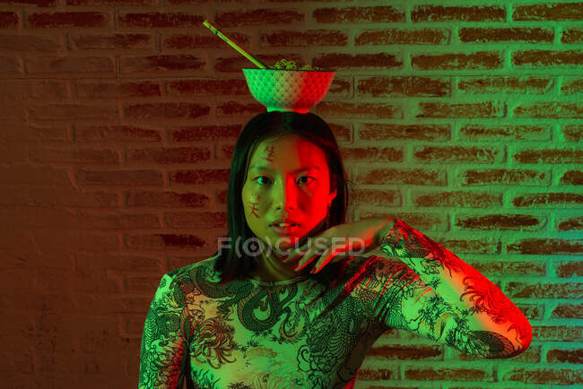 Emozionante giovane donna asiatica con geroglifici dipinti sul viso indossando abbigliamento elegante con ciotola di tagliatelle sulla testa toccando delicatamente il mento e guardando la fotocamera nella stanza buia — Foto stock