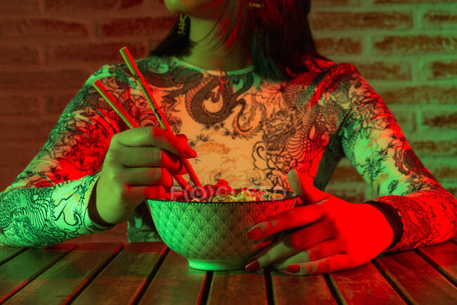 Неэмоциональная молодая азиатка с иероглифами на лице в стильной шляпе, поедающая вкусную лапшу, сидя в темной комнате напротив кирпичной стены — стоковое фото