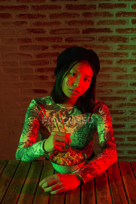 Jovem asiática sem emoção com hieróglifos pintados no rosto usando chapéu elegante comendo macarrão saboroso e olhando para a câmera enquanto sentado no quarto escuro contra a parede de tijolo — Fotografia de Stock