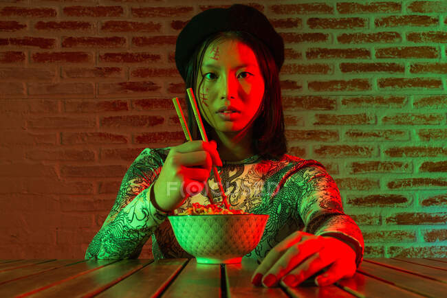 Unemotional young Asian female with hieroglyphs painted on face wearing stylish hat eating tasty noodle and looking at camera while sitting in dark room against brick wall — Stock Photo