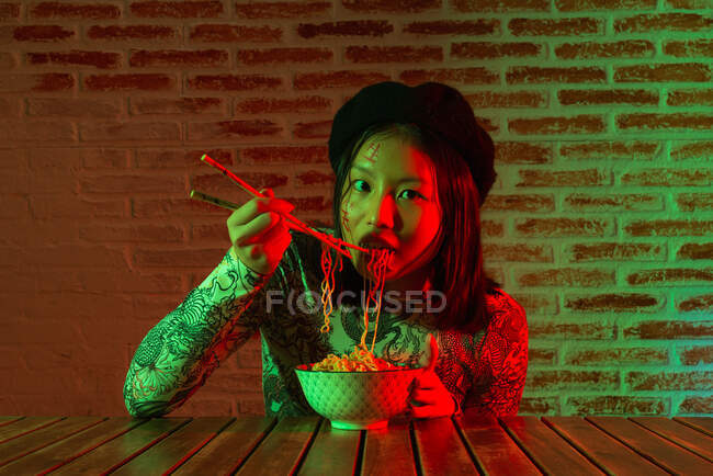 Unemotional young Asian female with hieroglyphs painted on face wearing stylish hat eating tasty noodle and looking at camera while sitting in dark room against brick wall — Stock Photo