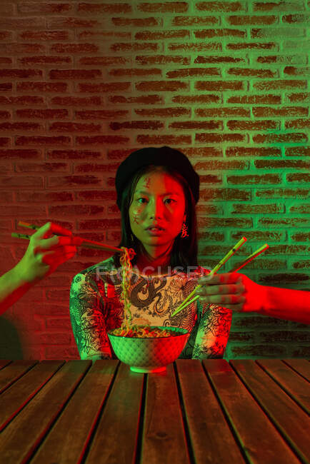 Crop anonymous people raising hands with chopsticks near amazed Asian woman with bowl with noodle — Stock Photo
