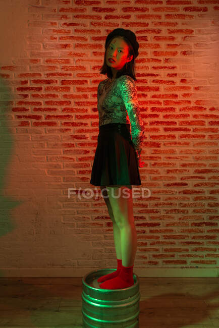 Corpo inteiro confiante bonita Asiática fêmea em mini-saia e chapéu elegante de pé graciosamente em barril de metal e olhando para a câmera durante sessão de fotos contra parede de tijolo em estúdio — Fotografia de Stock