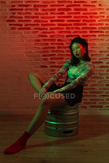 Corpo inteiro confiante bonita Asiática fêmea em mini-saia e chapéu elegante sentado graciosamente em barril de metal e olhando para a câmera durante sessão de fotos contra parede de tijolo em estúdio — Fotografia de Stock