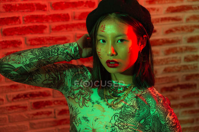 Unemotional giovane donna asiatica in elegante usura con geroglifici dipinti sul viso guardando la fotocamera con calma contro muro di mattoni in studio — Foto stock
