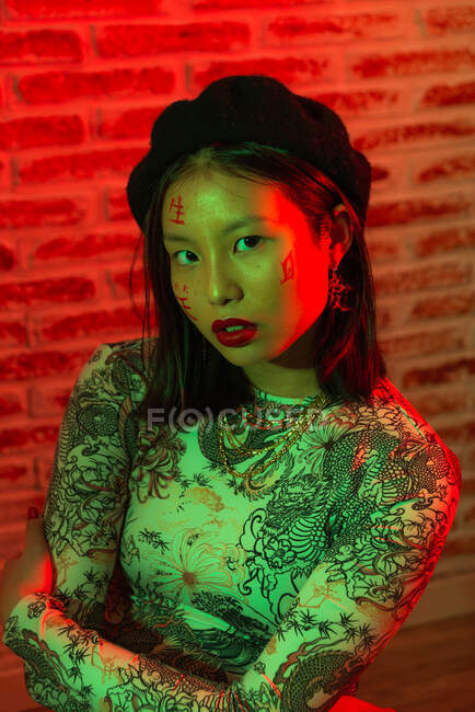 Unemotional young Asian female in stylish wear with hieroglyphs painted on face looking at camera calmly against brick wall in studio — Stock Photo