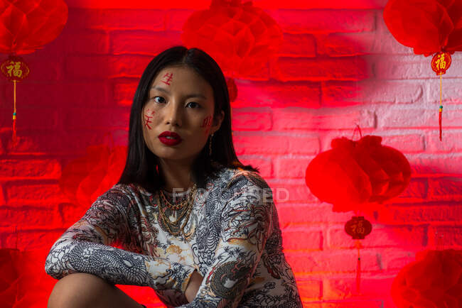 Unemotional giovane donna asiatica in elegante usura con geroglifici dipinti sul viso guardando la fotocamera con calma contro muro di mattoni in studio — Foto stock