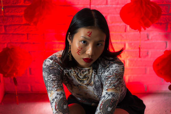 Joven hembra asiática sin emociones en ropa elegante con jeroglíficos pintados en la cara mirando a la cámara con calma contra la pared de ladrillo en el estudio - foto de stock