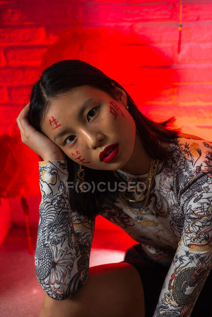 Atractiva mujer asiática de moda con jeroglíficos pintados en la cara con un atuendo elegante de pie con confianza y tocando la cara tiernamente mientras mira a la cámara en el estudio moderno - foto de stock