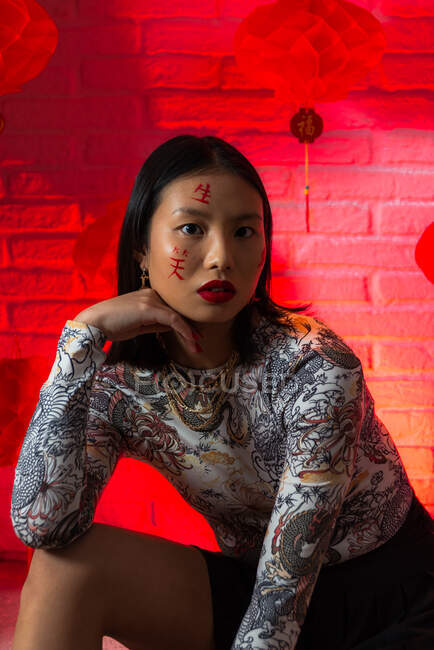 Atraente na moda asiática fêmea com hieróglifos pintados no rosto vestindo roupas elegantes sentado com confiança e tocando o rosto ternamente enquanto olha para a câmera no estúdio moderno — Fotografia de Stock