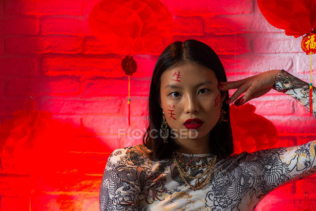 Atractiva mujer asiática de moda con jeroglíficos pintados en la cara con un atuendo elegante sentado con confianza y tocando la cara tiernamente mientras mira la cámara en el estudio moderno - foto de stock