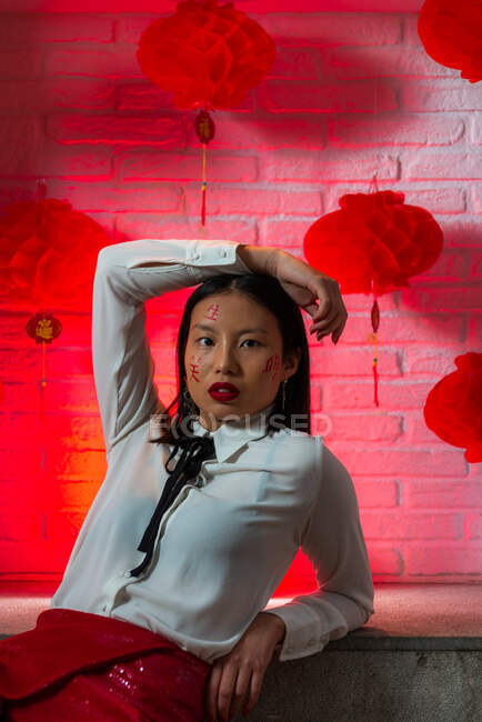 Belle femme asiatique confiante avec des hiéroglyphes peints sur le visage portant une mini-jupe rouge assise sur le sol et regardant la caméra pendant la séance photo contre un mur de briques en studio — Photo de stock