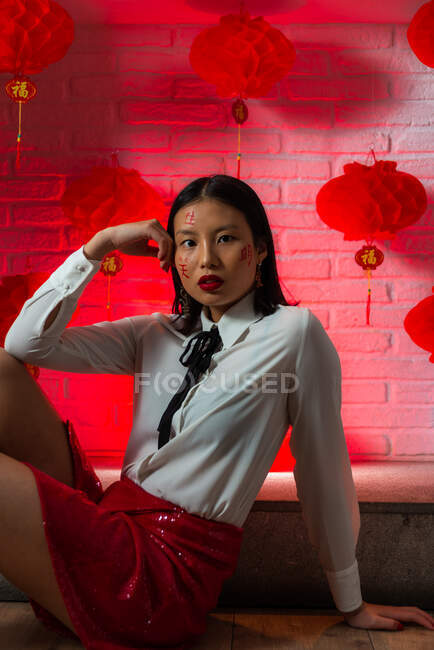 Mulher asiática confiante atraente com hieróglifos pintados no rosto vestindo mini-saia vermelha sentada no chão e olhando para a câmera durante sessão de fotos contra parede de tijolos em estúdio — Fotografia de Stock
