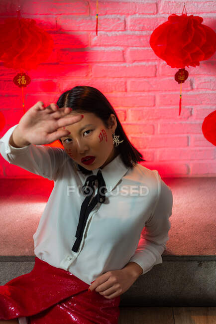 Belle femme asiatique confiante avec des hiéroglyphes peints sur le visage portant une mini-jupe rouge assise sur le sol et regardant la caméra pendant la séance photo contre un mur de briques en studio — Photo de stock
