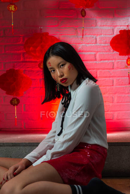Vista lateral atractiva mujer asiática segura con jeroglíficos pintados en la cara con mini falda roja sentada en el suelo y mirando a la cámara durante la sesión de fotos contra la pared de ladrillo en el estudio - foto de stock
