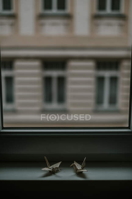 Origami di carta fatti a mano che rappresentano gru simili contro finestra e facciata della casa in crepuscolo su sfondo sfocato — Foto stock