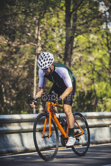 Full body of young sportsman in activewear and helmet riding bicycle on asphalt road amidst lush green trees on sunny day — Stock Photo