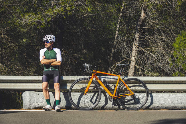 Full body of young male cyclist in protective helmet and sportswear standing leaning on crash barrier near bike parked on asphalt road against blue sky — Stock Photo