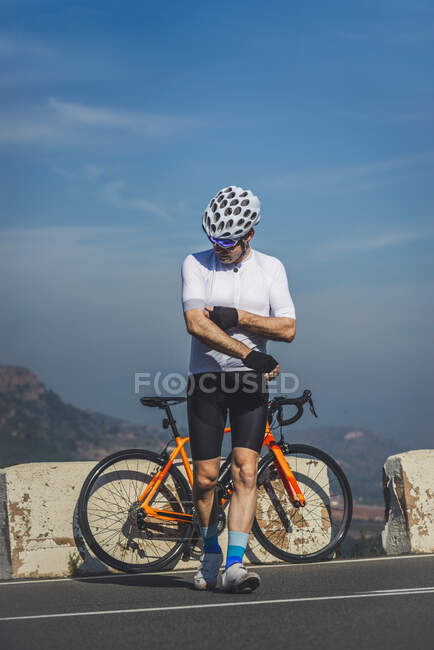 Повне тіло молодого чоловіка-велосипедиста в захисному шоломі та спортивному одязі, що йде біля велосипеда, припаркованого на асфальтній дорозі на блакитне небо — стокове фото
