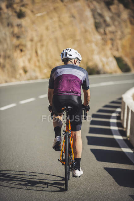 Back view of young sportsman in activewear and helmet riding bicycle on asphalt road on sunny day — Stock Photo