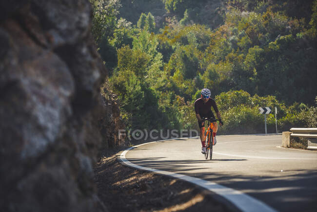 Corpo completo di giovane sportivo in activewear e casco in bicicletta su strada asfaltata sinuosa tra lussureggianti alberi verdi nella giornata di sole — Foto stock