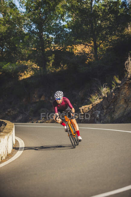 Full body of young sportsman in activewear and helmet riding bicycle on curvy asphalt road amidst lush green trees on sunny day — Stock Photo