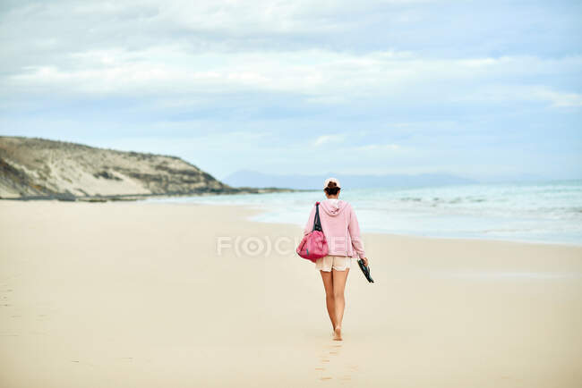 Voltar ver corpo inteiro de turista feminino anônimo com saco andando descalço ao longo da costa vazia em direção ao mar ondulando — Fotografia de Stock