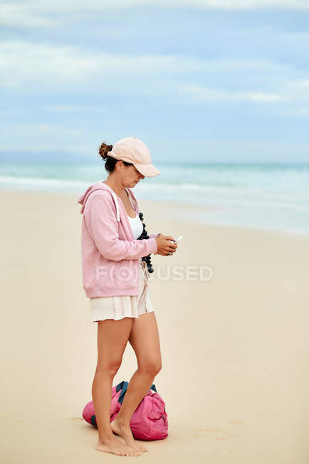 Боковой вид всего тела женщины-блогера-путешественника, просматривающего на смартфоне, стоя на песчаном пляже — стоковое фото