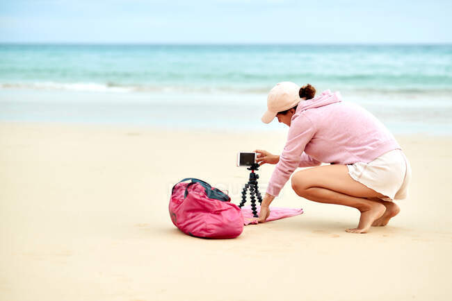 Visão lateral corpo inteiro do blogueiro de viagens feminino colocando celular no tripé na areia para filmar vídeo para as mídias sociais — Fotografia de Stock