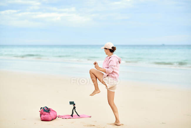 Visão lateral corpo inteiro do viajante feminino ativo levantando joelho durante o treinamento na praia de areia e gravação de vídeo no telefone móvel — Fotografia de Stock