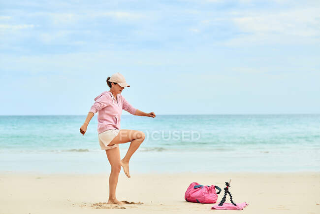 Side view full body of active female traveler lifting knee during training on sandy beach and recording video on mobile phone — Stock Photo