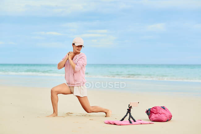 Side view full body of active female traveler doing lunges during training on sandy beach and recording video on mobile phone — Stock Photo