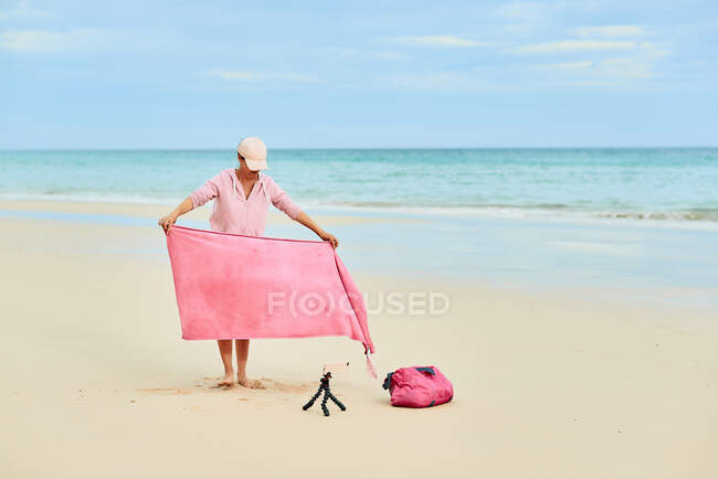 Full body of female tourist spreading towel on sandy coast near smartphone on tripod shooting video — Stock Photo