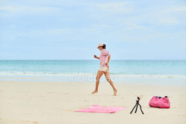 Seitenansicht Ganzkörper einer aktiven Reisenden, die während des Trainings am Sandstrand läuft und Videos auf dem Handy aufzeichnet — Stockfoto