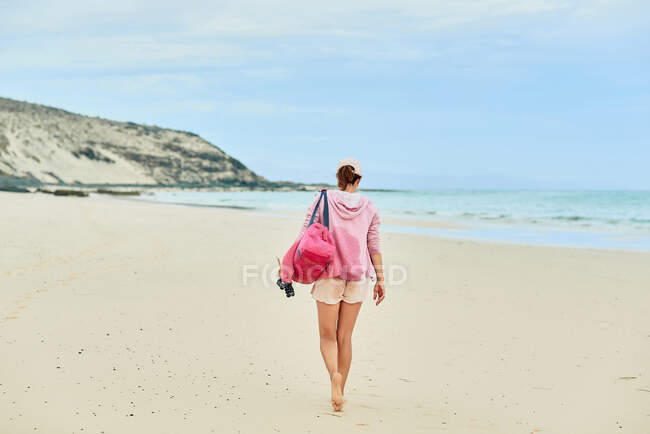 Задний вид полное тело анонимной туристки с мешком ходить босиком вдоль пустого побережья к машущему морю — стоковое фото