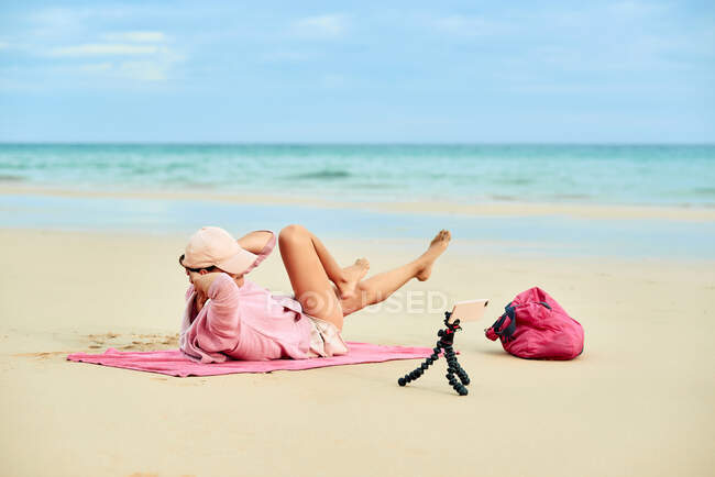 Vista lateral cuerpo completo irreconocible viajero femenino activo cruje ejercicio durante el entrenamiento en la playa de arena y la grabación de vídeo en el teléfono móvil - foto de stock